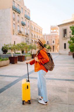 Sarı valizli, sırt çantalı ve güneşli meydanda yürüyen kadın bir gezginin dikiz görüntüsü. Güzel bir kadın küçük bir bavulla eski kasabanın sokaklarında dolaşıyor. Konsept: seyahat, turizm.
