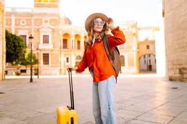 Sarı valizli, sırt çantalı ve güneşli meydanda yürüyen kadın bir gezginin dikiz görüntüsü. Güzel bir kadın küçük bir bavulla eski kasabanın sokaklarında dolaşıyor. Konsept: seyahat, turizm.