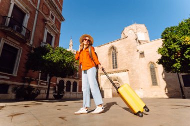 Sarı valizli, sırt çantalı ve güneşli meydanda yürüyen kadın bir gezginin dikiz görüntüsü. Güzel bir kadın küçük bir bavulla eski kasabanın sokaklarında dolaşıyor. Konsept: seyahat, turizm.