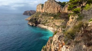 Parlak turkuaz suyu olan tenha bir plajın üzerinde görkemli uçurumlar yükseliyor. Güneş ufukta batıyor, gökyüzünde sıcak renkler bırakıyor ve sakin denizi yansıtıyor..