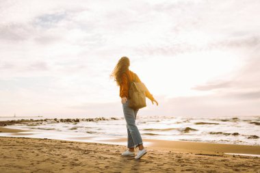 Sırt çantalı genç bir kadın kendini özgür hissediyor ve gün batımında deniz manzarasının tadını çıkarıyor. Güzel bir gezgin sahilde geziniyor. Özgürlük ve rahatlama kavramı.