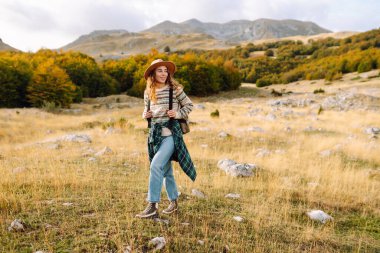 Şapka takan ve sırt çantası taşıyan yürüyüşçü dağlarda yürüyüş yolu boyunca yürür. Güzel bir kadın dağ manzarasından zevk alır ve gün batımında özgür hisseder. Seyahat ve yürüyüş kavramları. Etkin yaşam biçimi.