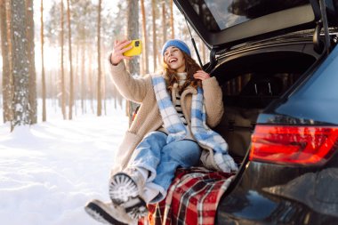 Gezgin bir telefon tutuyor, bir arabanın bagajında oturuyor, karlı ormanın tadını çıkarıyor. Telefon blogları olan ve yolculuk sırasında selfie çeken güzel bir kadın. Blogculuk, macera ve özgürlük kavramı.
