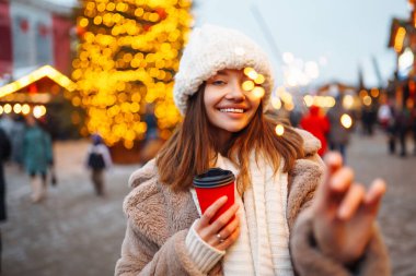 Mutlu bir kadın elinde parlak bir bardak sıcak içecekle Noel pazarında tatil ışıklarına karşı eğleniyor. Kadınlar karlı bir gün ve yeni yıl atmosferinin tadını çıkarırlar. Tatil kavramı.