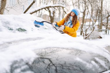 Parlak ceketli genç bir kadın güneşli ve karlı bir günde arabadan kar temizliyor. Özel bir fırçası olan bir kadın kışın bir arabadan karı temizler. Ulaşım konsepti, mevsimlik.