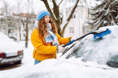 Parlak ceketli genç bir kadın güneşli ve karlı bir günde arabadan kar temizliyor. Özel bir fırçası olan bir kadın kışın bir arabadan karı temizler. Ulaşım konsepti, mevsimlik.