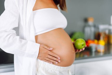 Gülümseyen hamile bir kadın, elinde yeşil bir elmayla şık bir mutfakta duruyor. Bebek bekleyen genç bir kadın, meyve yiyor ve sıcak bir odada vakit geçiriyor. Konsept gıda, sağlık ve rahatlıkla ilgili..