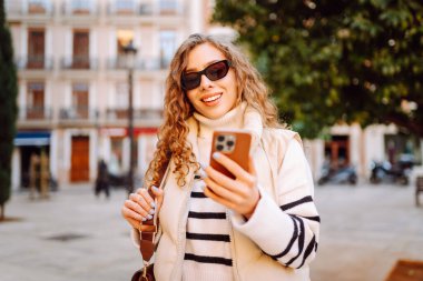 Güneş gözlüklü genç bir kadın, elinde telefon, güneşli bir günde şehrin eski sokaklarında geziniyor. Güzel bir turist gün batımını, blogları ve telefondaki mesajları sever. Turizm kavramı, hafta sonları.