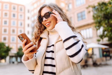 Güneş gözlüklü genç bir kadın, elinde telefon, güneşli bir günde şehrin eski sokaklarında geziniyor. Güzel bir turist gün batımını, blogları ve telefondaki mesajları sever. Turizm kavramı, hafta sonları.