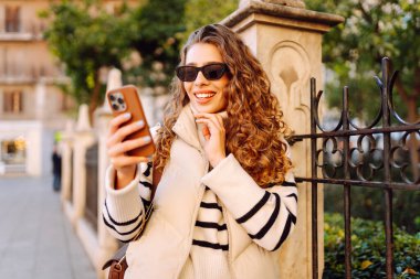 Güneş gözlüklü genç bir kadın, elinde telefon, güneşli bir günde şehrin eski sokaklarında geziniyor. Güzel bir turist gün batımını, blogları ve telefondaki mesajları sever. Turizm kavramı, hafta sonları.