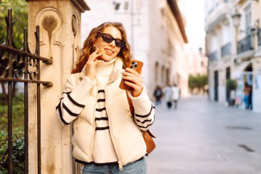 Güneş gözlüklü genç bir kadın, elinde telefon, güneşli bir günde şehrin eski sokaklarında geziniyor. Güzel bir turist gün batımını, blogları ve telefondaki mesajları sever. Turizm kavramı, hafta sonları.