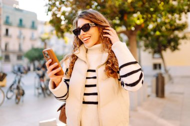 Güneş gözlüklü genç bir kadın, elinde telefon, güneşli bir günde şehrin eski sokaklarında geziniyor. Güzel bir turist gün batımını, blogları ve telefondaki mesajları sever. Turizm kavramı, hafta sonları.