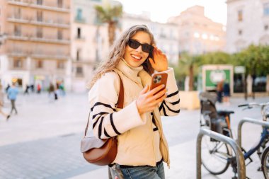 Güneş gözlüklü genç bir kadın, elinde telefon, güneşli bir günde şehrin eski sokaklarında geziniyor. Güzel bir turist gün batımını, blogları ve telefondaki mesajları sever. Turizm kavramı, hafta sonları.