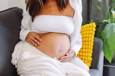 Hamile bir kadın rahat bir odada kanepede oturur ve elleriyle karnına hafifçe dokunur. Bebek bekleyen güzel bir kadın güneşli bir oturma odasında dinleniyor. Beklenti ve rahatlama kavramı.