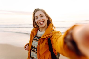 Sırt çantası ve telefonu olan neşeli bir kadın gün batımında sahilde selfie çekiyor. Genç bir gezgin telefonunda blog yazıyor ve deniz kenarında güneşli bir günün tadını çıkarıyor. Seyahat ve günlük tutma kavramı.