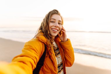 Sırt çantası ve telefonu olan neşeli bir kadın gün batımında sahilde selfie çekiyor. Genç bir gezgin telefonunda blog yazıyor ve deniz kenarında güneşli bir günün tadını çıkarıyor. Seyahat ve günlük tutma kavramı.