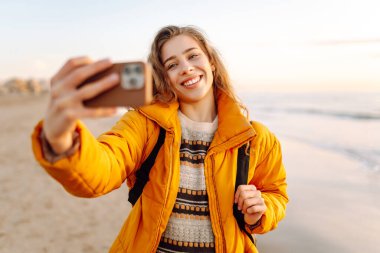 Sırt çantası ve telefonu olan neşeli bir kadın gün batımında sahilde selfie çekiyor. Genç bir gezgin telefonunda blog yazıyor ve deniz kenarında güneşli bir günün tadını çıkarıyor. Seyahat ve günlük tutma kavramı.