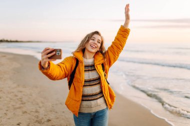 Sırt çantası ve telefonu olan neşeli bir kadın gün batımında sahilde selfie çekiyor. Genç bir gezgin telefonunda blog yazıyor ve deniz kenarında güneşli bir günün tadını çıkarıyor. Seyahat ve günlük tutma kavramı.
