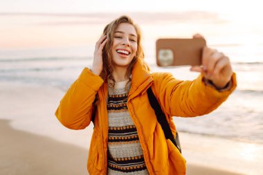 Sırt çantası ve telefonu olan neşeli bir kadın gün batımında sahilde selfie çekiyor. Genç bir gezgin telefonunda blog yazıyor ve deniz kenarında güneşli bir günün tadını çıkarıyor. Seyahat ve günlük tutma kavramı.