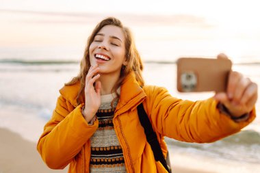 Sırt çantası ve telefonu olan neşeli bir kadın gün batımında sahilde selfie çekiyor. Genç bir gezgin telefonunda blog yazıyor ve deniz kenarında güneşli bir günün tadını çıkarıyor. Seyahat ve günlük tutma kavramı.