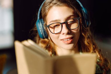 Kablosuz kulaklık takan ve rahat bir odada kitap okuyan genç bir kadının yakın çekimi. Güzel bir kadın kitap okumayı ve samimi atmosferde müzik dinlemeyi sever. Rahatlama ve keyif kavramı.
