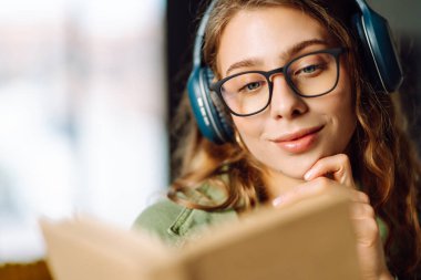 Kablosuz kulaklık takan ve rahat bir odada kitap okuyan genç bir kadının yakın çekimi. Güzel bir kadın kitap okumayı ve samimi atmosferde müzik dinlemeyi sever. Rahatlama ve keyif kavramı.