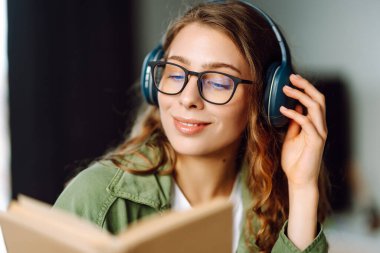 Kablosuz kulaklık takan ve rahat bir odada kitap okuyan genç bir kadının yakın çekimi. Güzel bir kadın kitap okumayı ve samimi atmosferde müzik dinlemeyi sever. Rahatlama ve keyif kavramı.