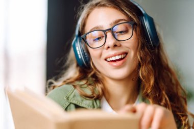 Kablosuz kulaklık takan ve rahat bir odada kitap okuyan genç bir kadının yakın çekimi. Güzel bir kadın kitap okumayı ve samimi atmosferde müzik dinlemeyi sever. Rahatlama ve keyif kavramı.
