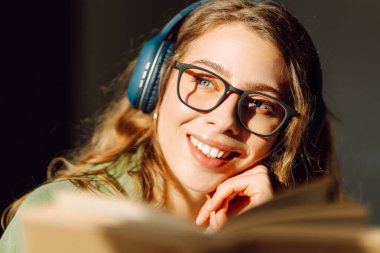 Kablosuz kulaklık takan ve rahat bir odada kitap okuyan genç bir kadının yakın çekimi. Güzel bir kadın kitap okumayı ve samimi atmosferde müzik dinlemeyi sever. Rahatlama ve keyif kavramı.