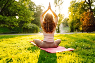 Spor elbiseli güzel bir kadın gün batımında yoga minderinde meditasyon yapıyor. Atletik bir kadın güneşli bir parkta yoga yapıyor ve doğanın tadını çıkarıyor. Yoga kavramları, esneme. Sağlıklı yaşam tarzı.