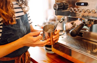 Deneyimli bir barista sıcak bir kafede taze demlenmiş kahve hazırlar. Genç bir kadın espressoya süt döker ve köpürür. Küçük iş konsepti, içecekler.
