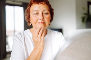 Yaşlı bir kadın nazikçe yüzüne dokunuyor, aynada tenini inceliyor. Altmışlarında gülümseyen bir kadın, rahat bir odada otururken güzelliğinin tadını çıkarır. Güzellik anlayışı, mutlu emeklilik.