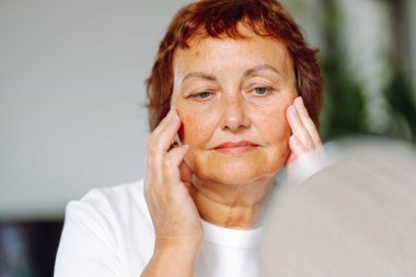 Yaşlı bir kadın nazikçe yüzüne dokunuyor, aynada tenini inceliyor. Altmışlarında gülümseyen bir kadın, rahat bir odada otururken güzelliğinin tadını çıkarır. Güzellik anlayışı, mutlu emeklilik.