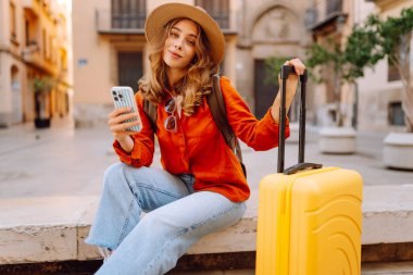 Şapkalı neşeli bayan turist ve telefon ile şehrin caddelerinde sarı valiz gezintileri yapıyor. Telefonu olan genç bir kadın güneşli bir günde blog yazıyor, gezinin tadını çıkarıyor. Bloglar, hafta sonları için kavramlar.