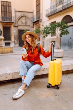 Şapkalı neşeli bayan turist ve telefon ile şehrin caddelerinde sarı valiz gezintileri yapıyor. Telefonu olan genç bir kadın güneşli bir günde blog yazıyor, gezinin tadını çıkarıyor. Bloglar, hafta sonları için kavramlar.