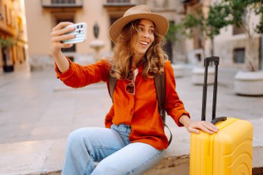 Şapkalı neşeli bayan turist ve telefon ile şehrin caddelerinde sarı valiz gezintileri yapıyor. Telefonu olan genç bir kadın güneşli bir günde blog yazıyor, gezinin tadını çıkarıyor. Bloglar, hafta sonları için kavramlar.