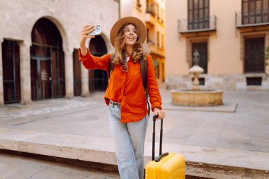Şapkalı neşeli bayan turist ve telefon ile şehrin caddelerinde sarı valiz gezintileri yapıyor. Telefonu olan genç bir kadın güneşli bir günde blog yazıyor, gezinin tadını çıkarıyor. Bloglar, hafta sonları için kavramlar.