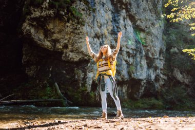 Parlak sırt çantalı bir yürüyüşçü ormandaki yüksek dağ nehri yakınlarında yürüyüş yolu boyunca yürür. Güzel bir kadın güneşli bir günde nehir ve dağ manzarası boyunca yürümekten hoşlanır. Konseptler: doğa, yürüyüş.
