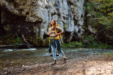 Parlak sırt çantalı bir yürüyüşçü ormandaki yüksek dağ nehri yakınlarında yürüyüş yolu boyunca yürür. Güzel bir kadın güneşli bir günde nehir ve dağ manzarası boyunca yürümekten hoşlanır. Konseptler: doğa, yürüyüş.