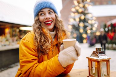 Sarı ceketli gülümseyen kadın, hareketli kış pazarında sıcak bir içecek tutuyor. Güzel turistler yılbaşı atmosferinin tadını çıkarıyorlar. Işıklarla süslenmiş, elinde sıcak şarapla. Kavram şenliktir, samimidir..