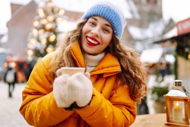 Sarı ceketli gülümseyen kadın, hareketli kış pazarında sıcak bir içecek tutuyor. Güzel turistler yılbaşı atmosferinin tadını çıkarıyorlar. Işıklarla süslenmiş, elinde sıcak şarapla. Kavram şenliktir, samimidir..