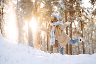 Mavi atkı ve şapkalı genç bir kadın güneşli bir günde karda eğleniyor. Güzel bir kadın havaya kar fırlatır ve ormanda kış gününün tadını çıkarır. Eğlence, rahatlama ve mevsimlik kavramları.