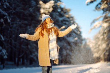 Mavi atkı ve şapkalı genç bir kadın güneşli bir günde karda eğleniyor. Güzel bir kadın havaya kar fırlatır ve ormanda kış gününün tadını çıkarır. Eğlence, rahatlama ve mevsimlik kavramları.