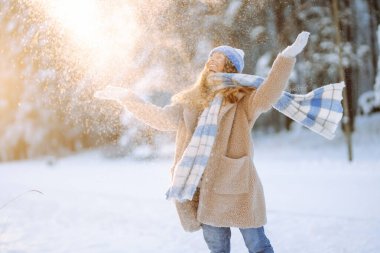 Mavi atkı ve şapkalı genç bir kadın güneşli bir günde karda eğleniyor. Güzel bir kadın havaya kar fırlatır ve ormanda kış gününün tadını çıkarır. Eğlence, rahatlama ve mevsimlik kavramları.