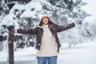 Sıcak kıyafetler giymiş neşeli bir kadın karlı bir ormanda eğleniyor. Güzel bir kadın kış gününün tadını çıkarır, havaya kar atar ve dışarıda dinlenir. Eğlence, hafta sonları ve don kavramları.
