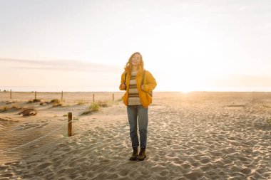 Parlak ceketli genç bir kadın gün batımında kumlu bir sahilde yürüyor. Güzel bir sırt çantalı doğa ve açık hava macerasından zevk alır. Seyahat, doğa ve rahatlama kavramı.