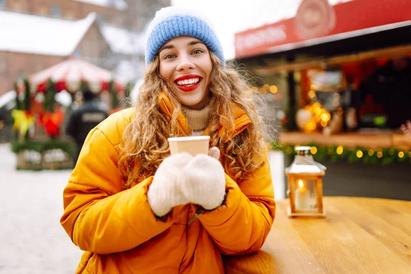 Sarı ceketli gülümseyen kadın, hareketli kış pazarında sıcak bir içecek tutuyor. Güzel turistler yılbaşı atmosferinin tadını çıkarıyorlar. Işıklarla süslenmiş, elinde sıcak şarapla. Kavram şenliktir, samimidir..