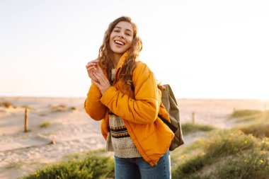 Parlak ceketli genç bir kadın gün batımında kumlu bir sahilde yürüyor. Güzel bir sırt çantalı doğa ve açık hava macerasından zevk alır. Seyahat, doğa ve rahatlama kavramı.