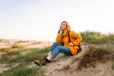 Parlak ceketli genç bir kadın gün batımında kumlu bir sahilde yürüyor. Güzel bir sırt çantalı doğa ve açık hava macerasından zevk alır. Seyahat, doğa ve rahatlama kavramı.