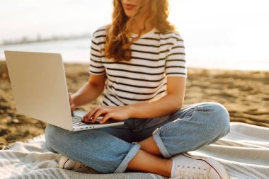 Güneşli bir günde plaj battaniyesinde otururken dizüstü bilgisayarla çalışan serbest çalışan bir kadın. Dizüstü bilgisayarı olan güzel bir kadın gün batımında doğanın ve deniz manzarasının tadını çıkarıyor. Rahatlama kavramı, dinlenme.